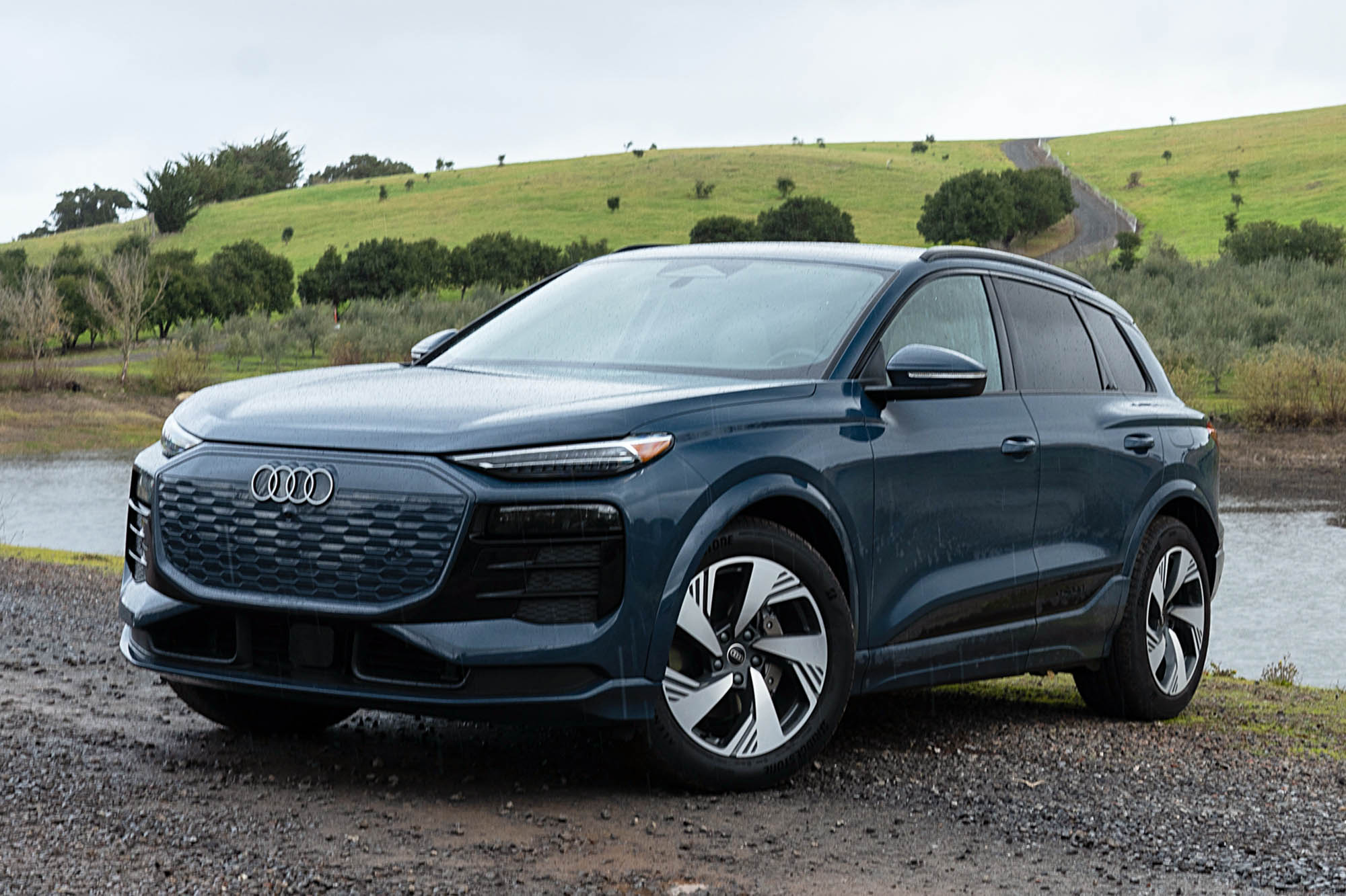 2025 Audi Q6 e-tron Prestige in Plasma Blue with a pond and green, rolling hillside in the background.