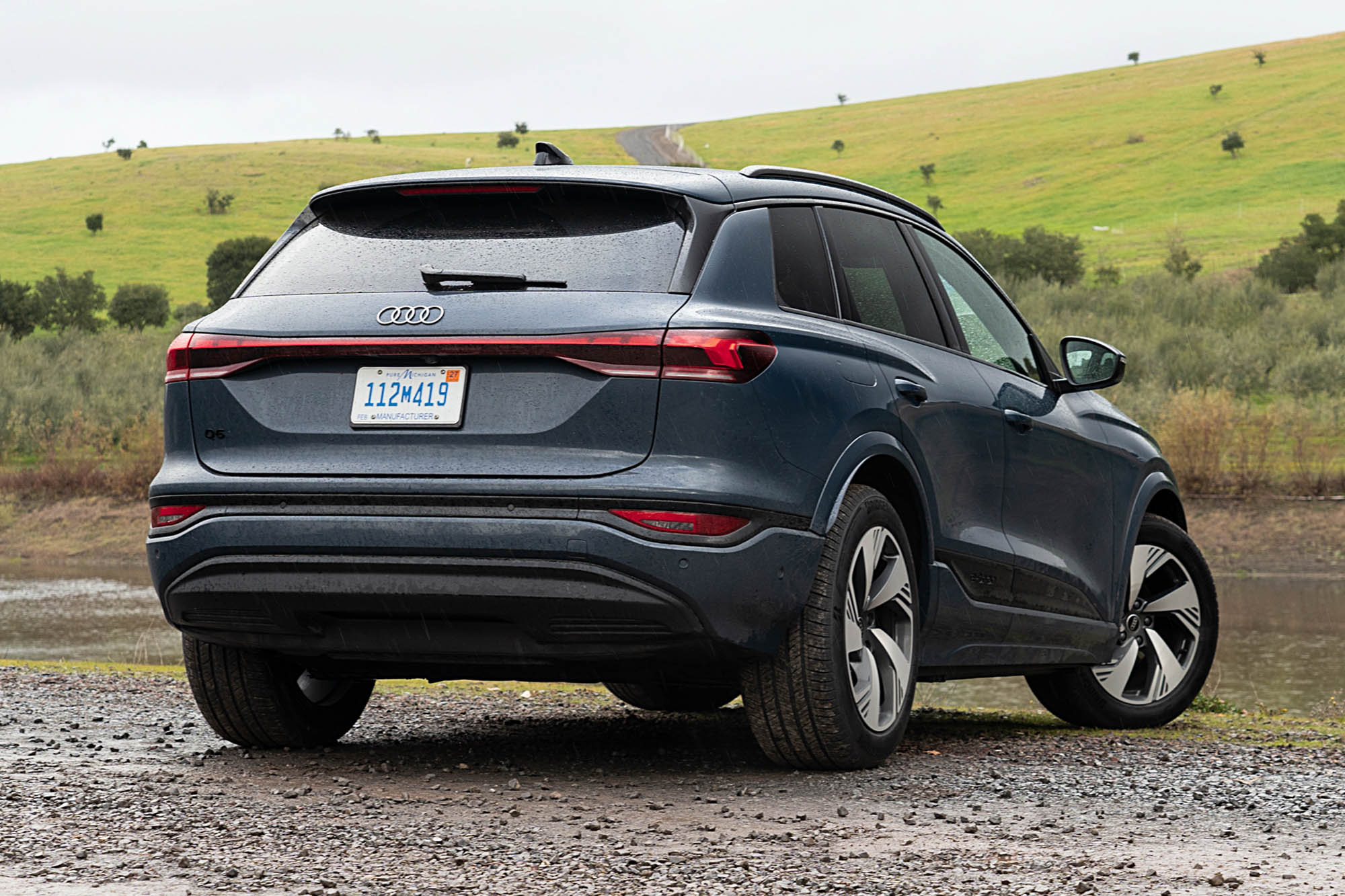 2025 Audi Q6 e-tron Prestige in Plasma Blue with a pond and green, rolling hillside in the background.