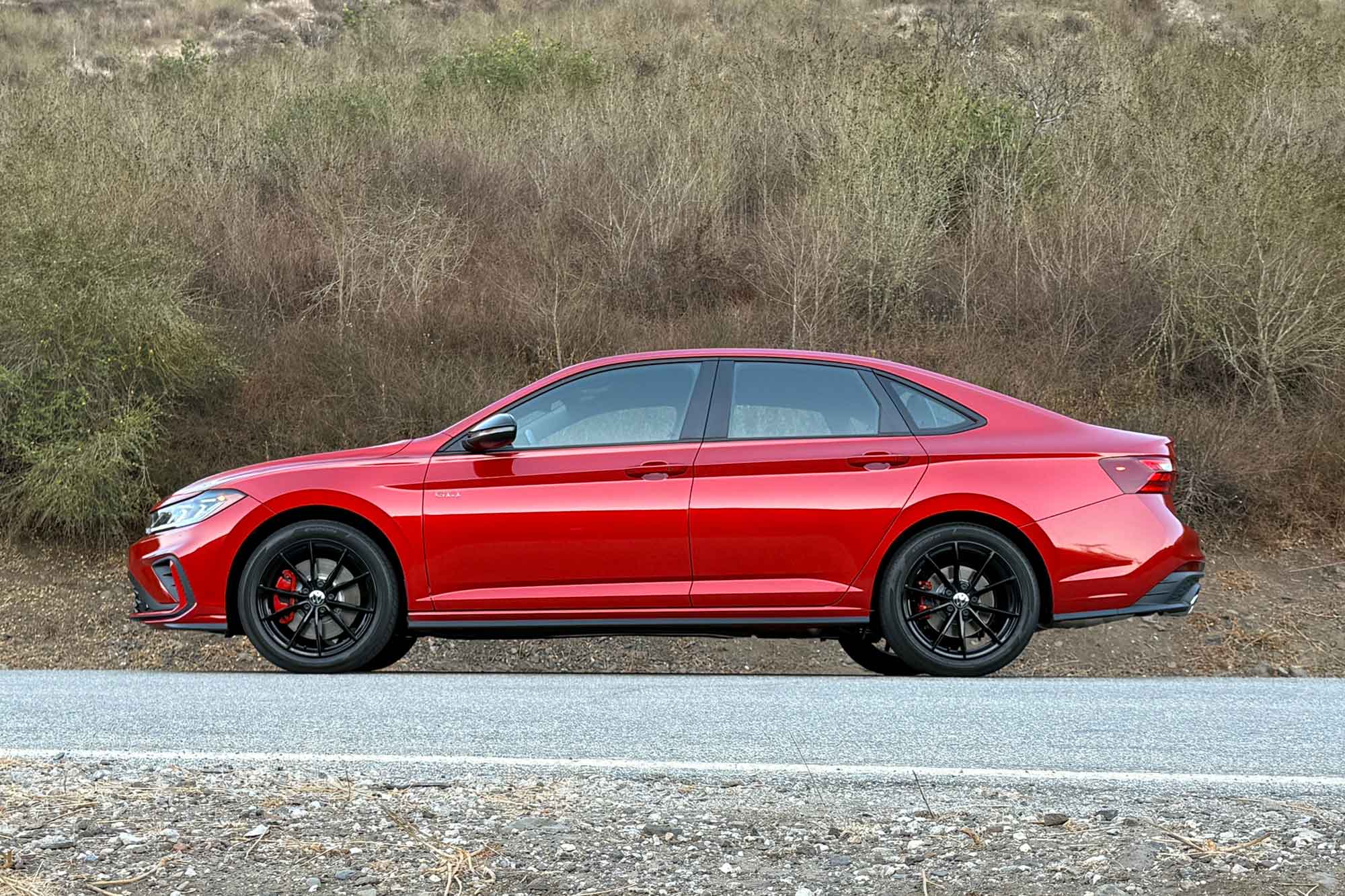 2025 Volkswagen Jetta GLI in Kings Red Metallic with Black package, shown with a brush-covered hillside in the background.