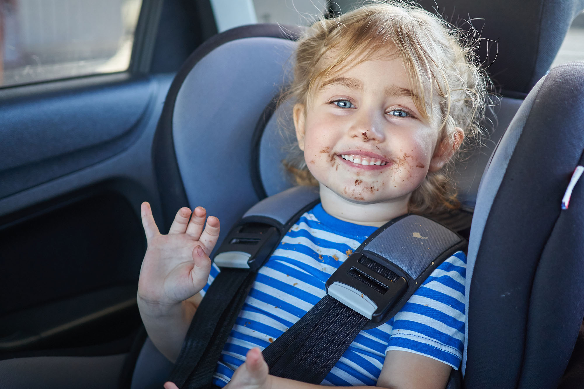 Child with crumbs and food on their body sits in car seat smiling at the camera