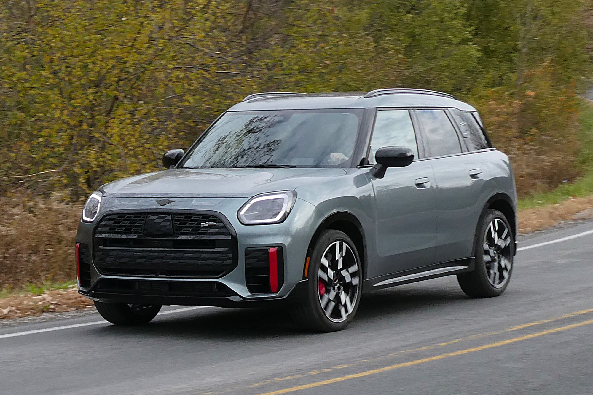 2025 Mini Countryman John Cooper Works in Smokey Green driving on the road with trees in the background.