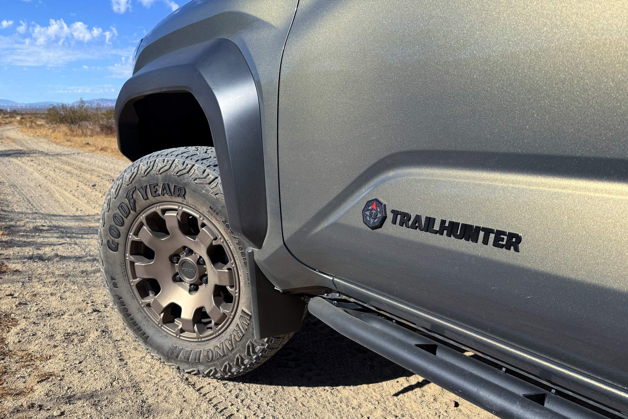 2024 Toyota Tacoma Trailhunter showing the door emblem, front wheel, rugged-terrain tire, and rock rails with a desert trail stretching into the distance with blue skies in the background