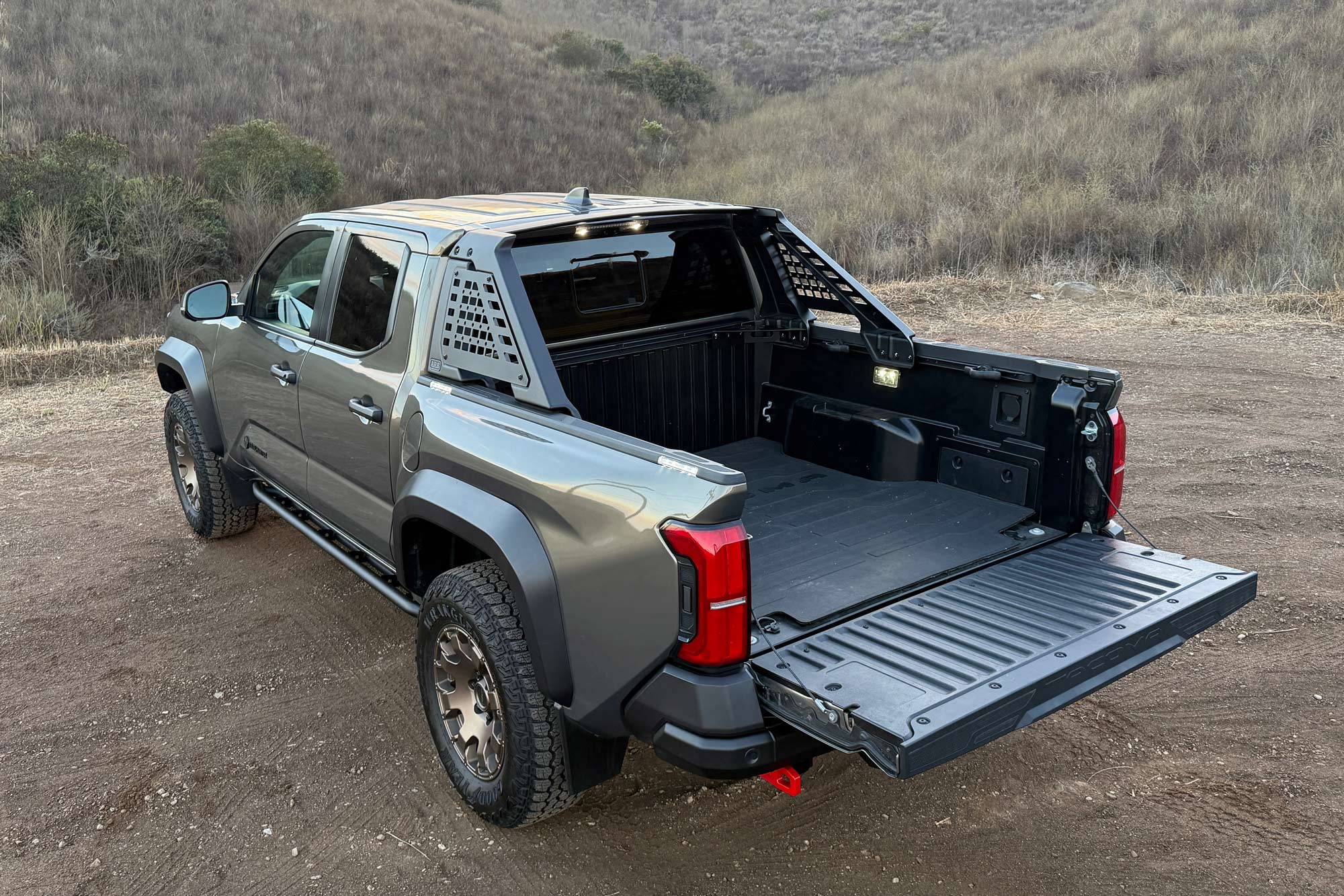 Rear quarter 2024 Toyota Tacoma Trailhunter in Bronze Oxide showing the cargo bed and LED scene lighting with a mountain valley in the background