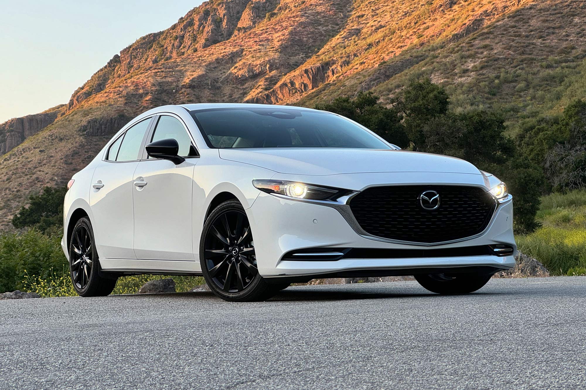 Front-quarter view of a white 2024 Mazda 3 2.5 Turbo Premium Plus parked on the pavement with a sunlit mountain in the background.