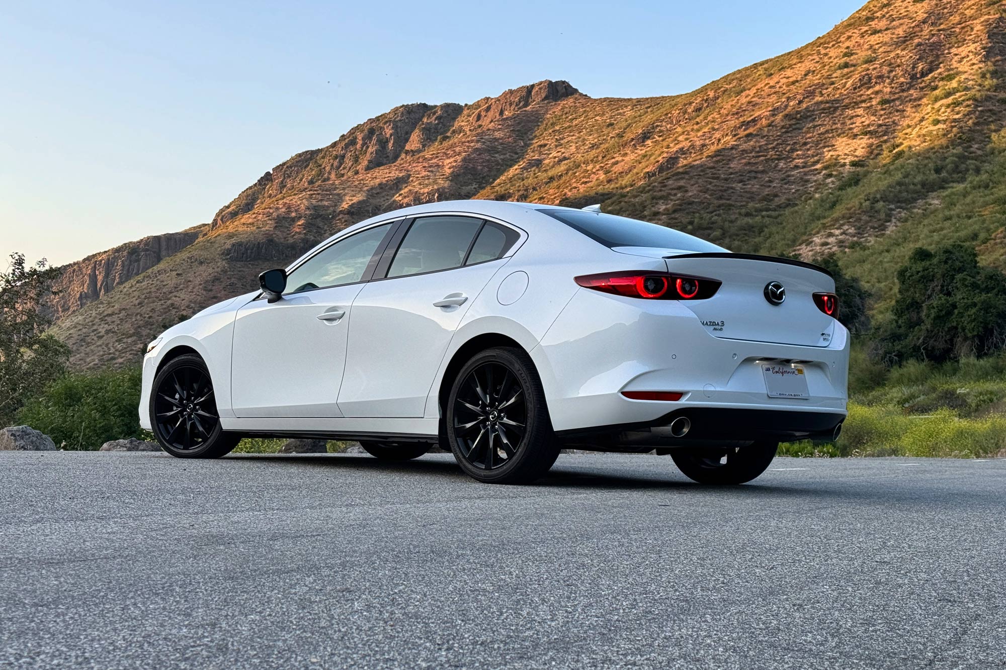 Rear-quarter view of a white 2024 Mazda 3 2.5 Turbo Premium Plus parked on the pavement with a sunlit mountain in the background.