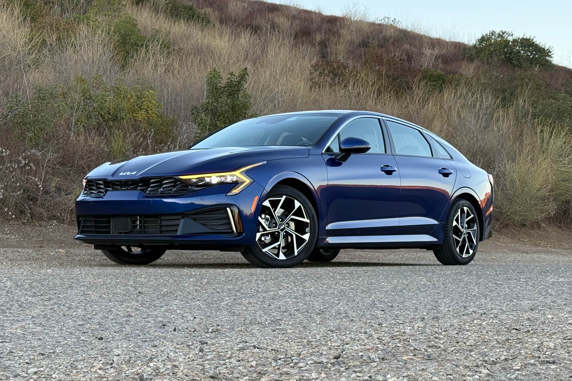 2025 Kia K5 EX in Yacht Blue with a brush-covered hillside in the background.