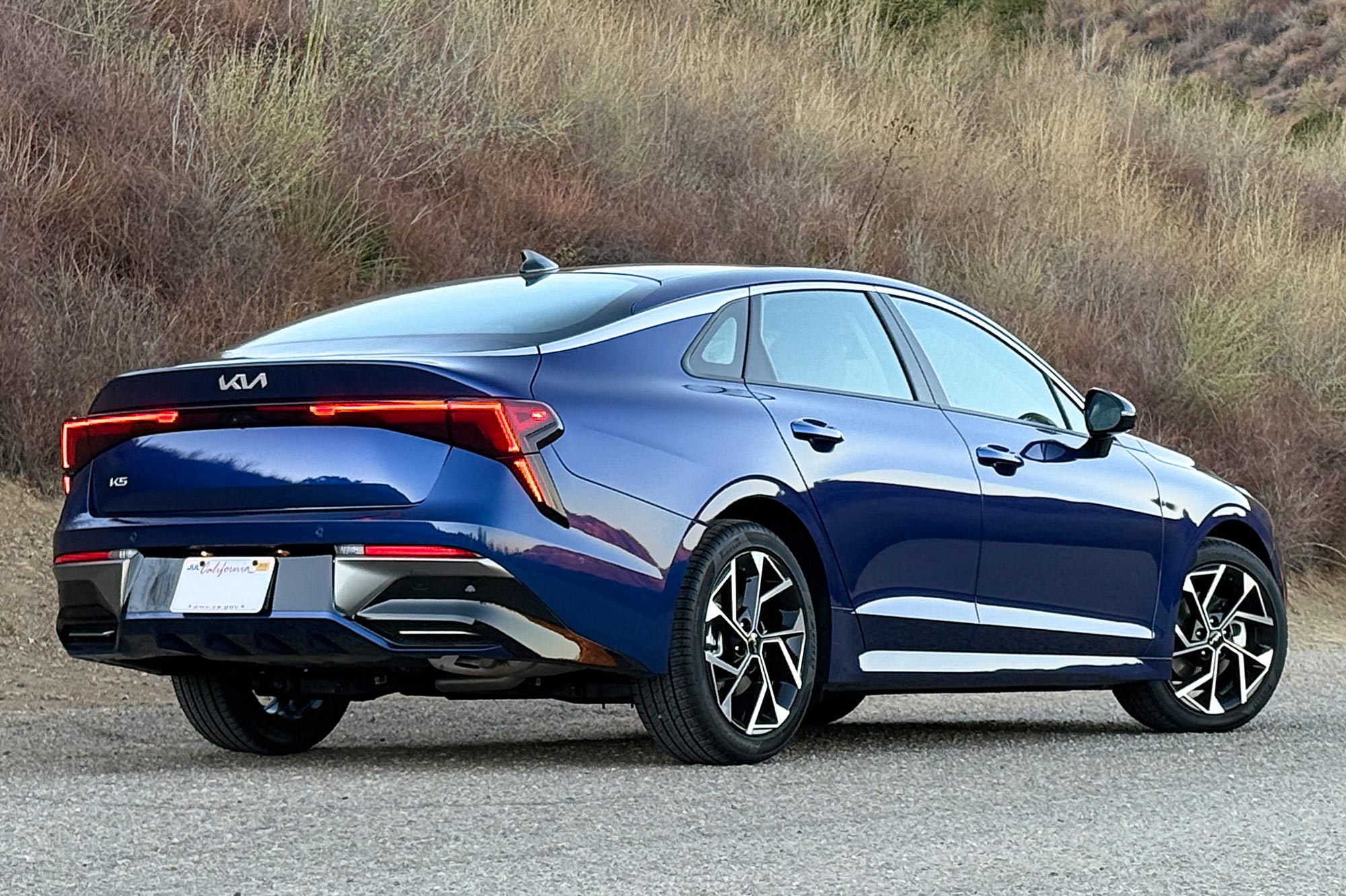 2025 Kia K5 EX in Yacht Blue with a brush-covered hillside in the background.