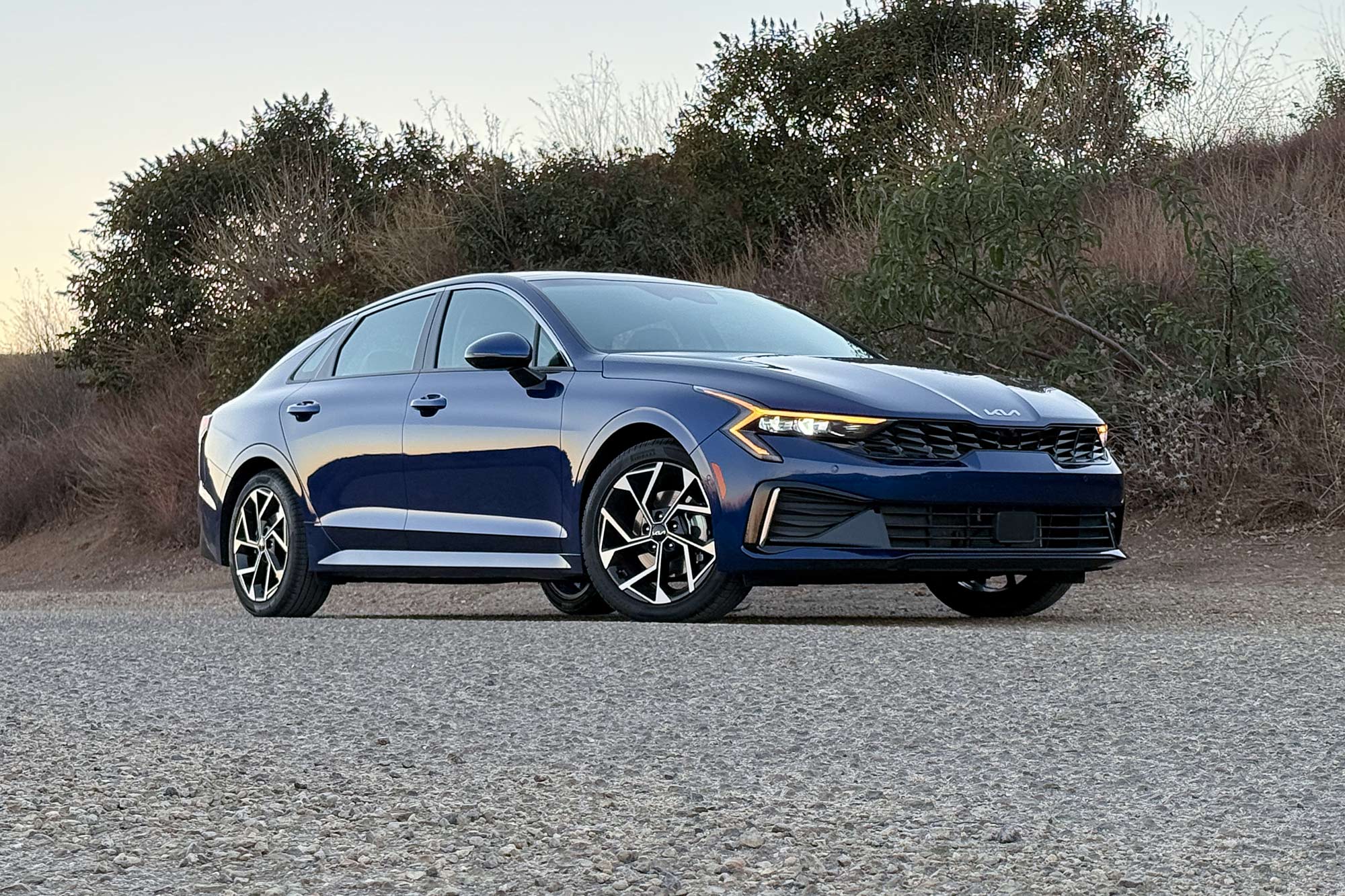 2025 Kia K5 EX in Yacht Blue with a brush-covered hillside in the background.