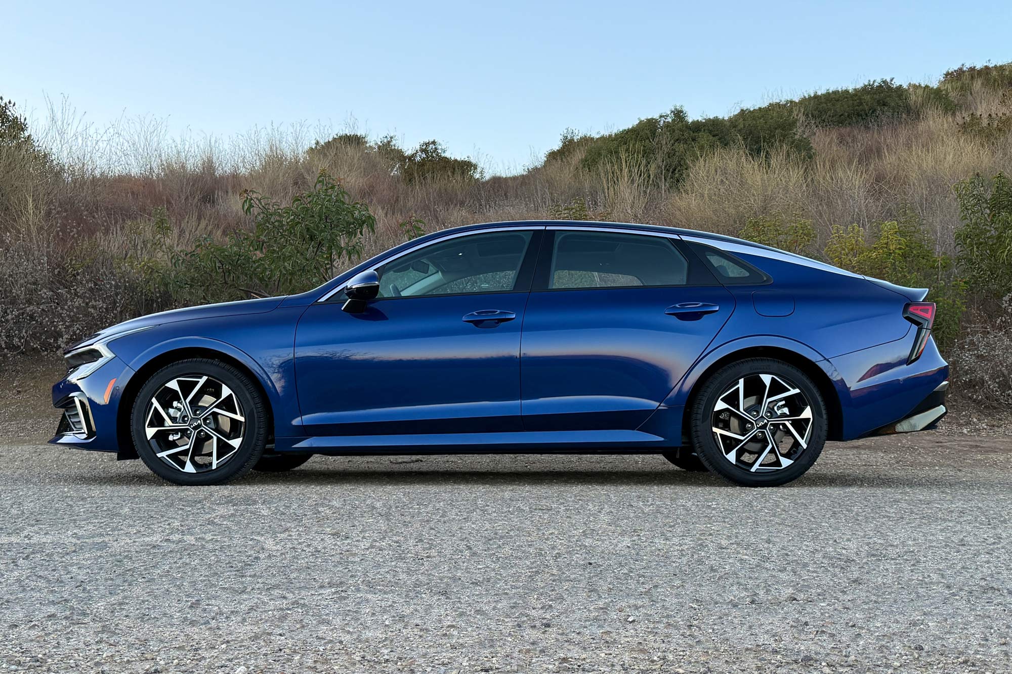 2025 Kia K5 EX in Yacht Blue with a brush-covered hillside in the background.